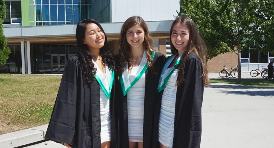 Tre studentesse della Oak Bay Secondary High School in toga da diploma stanno davanti a un edificio universitario con biciclette e un'area verde.