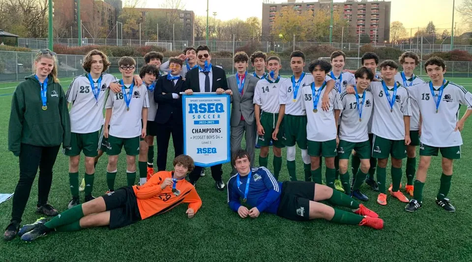 Un gruppo di studenti della St. Thomas High School festeggia la vittoria sul campo da calcio con coppa e medaglie davanti allo sfondo della città.