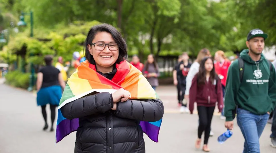 Una persona sorridente con una sciarpa arcobaleno colorata si trova su un viale alberato nel campus della California State University Long Beach.