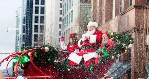 Papá Noel y dos niños disfrazados de elfos saludan desde un trineo rojo decorado con guirnaldas