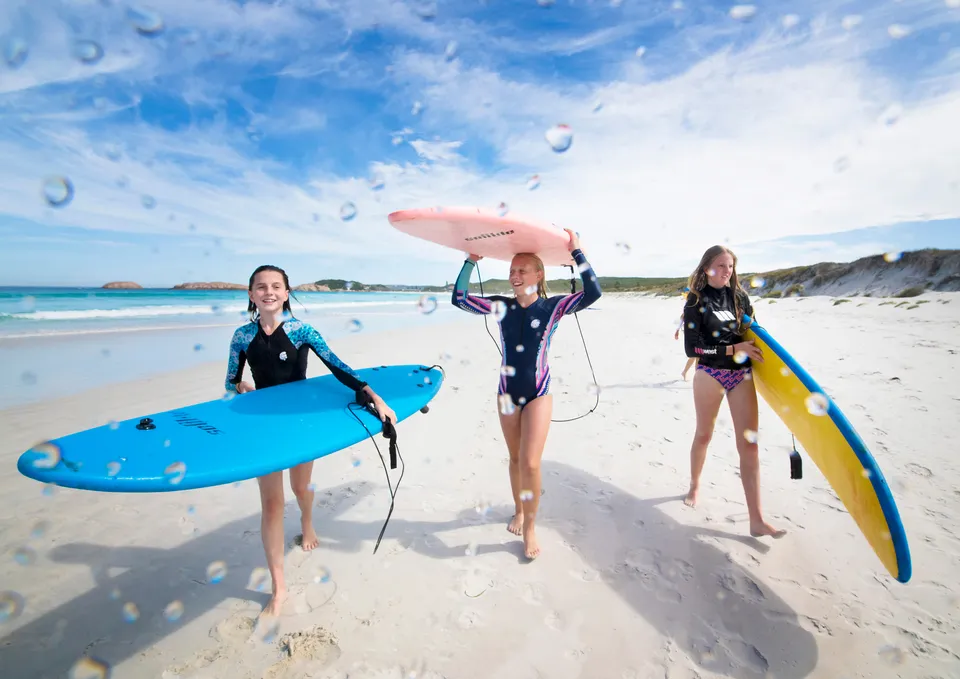 Tre studentesse della Esperance Anglican Community School camminano con tavole da surf su una spiaggia di sabbia.