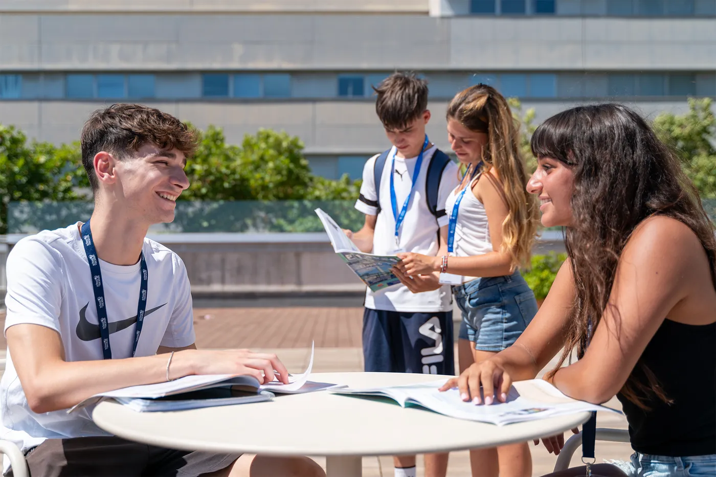 Studenti sorridenti con lanyard studiano insieme attorno a un tavolo nel campus soleggiato di una vacanza studio