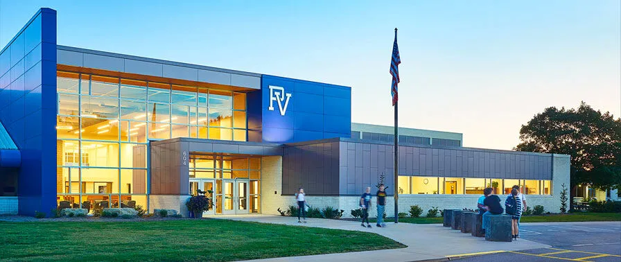 El moderno edificio de varios pisos de la Pleasant Valley Secondary School con fachada azul y grandes ventanas está rodeado por un césped y personas frente a la entrada.