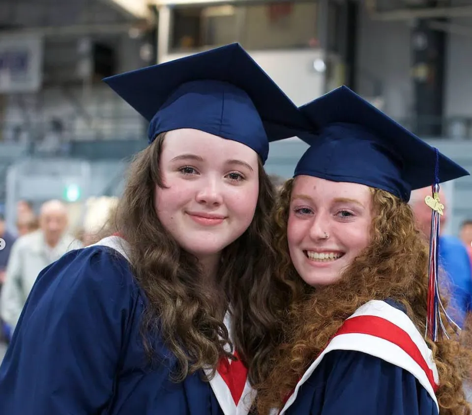 Due studentesse in abito da diploma sorridono e posano insieme davanti a uno sfondo sfocato nel campus della Timberline Secondary School.