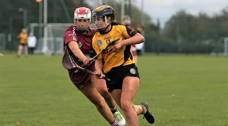 Due studentesse del Duiske College giocano a un gioco sportivo su un campo in erba circondato da alberi.