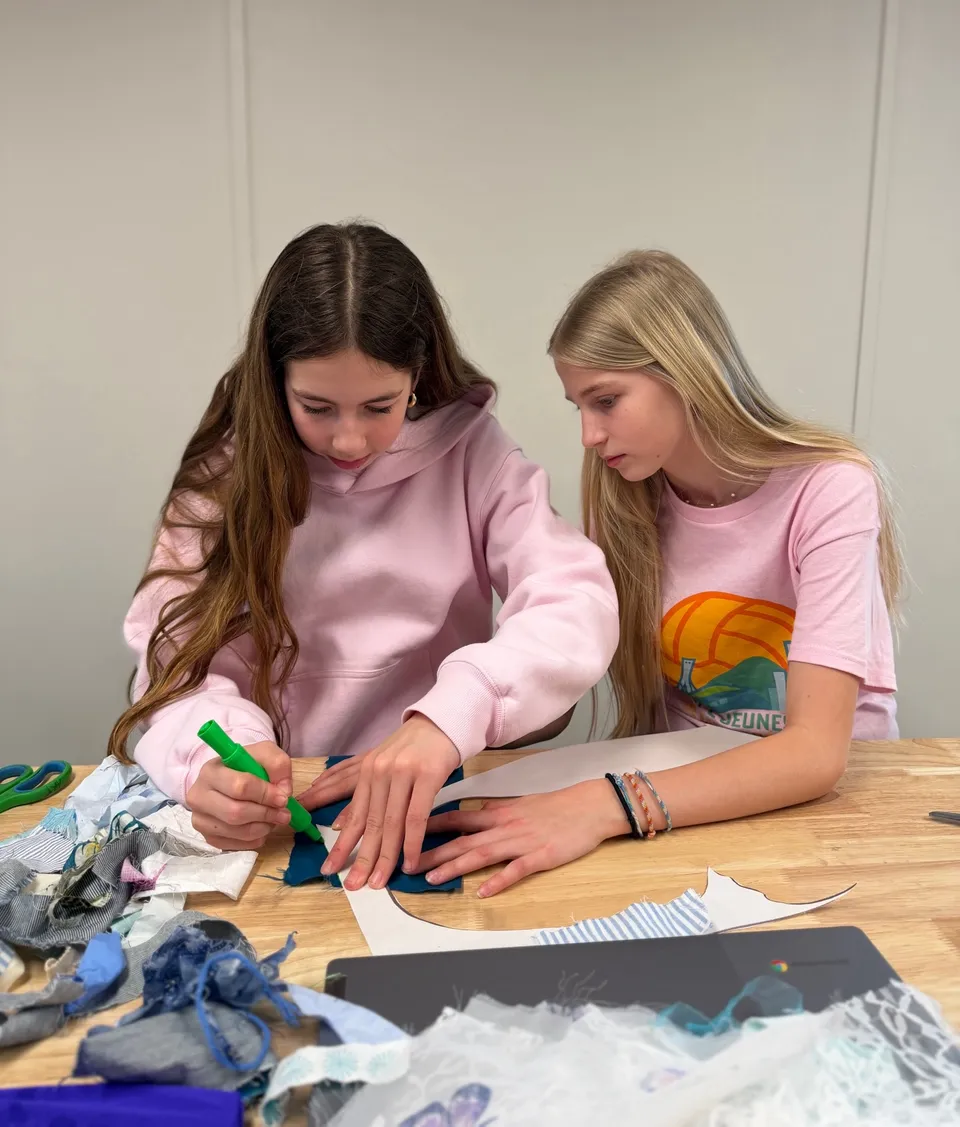 Due studentesse della Aspengrove School lavorano insieme a un progetto tessile con stoffe su un tavolo di legno.