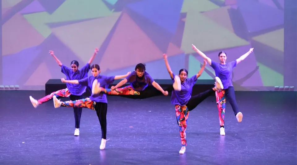 Un gruppo di studenti della Girrawheen Senior High School balla sul palco davanti a uno sfondo di forme geometriche viola e blu.