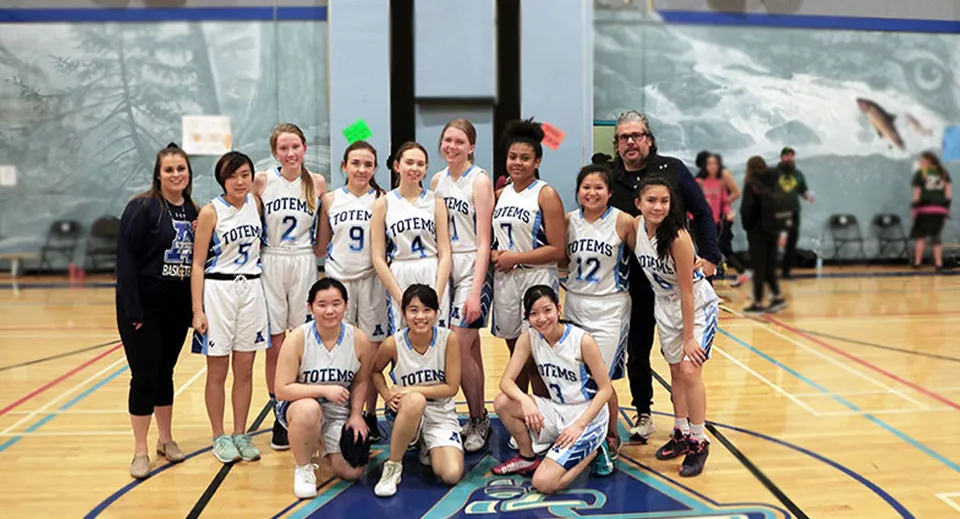 Un gruppo di giovani studentesse della Aldergrove Community Secondary School posa in divisa da basket sul campo da gioco davanti a una parete di vetro con un paesaggio innevato.