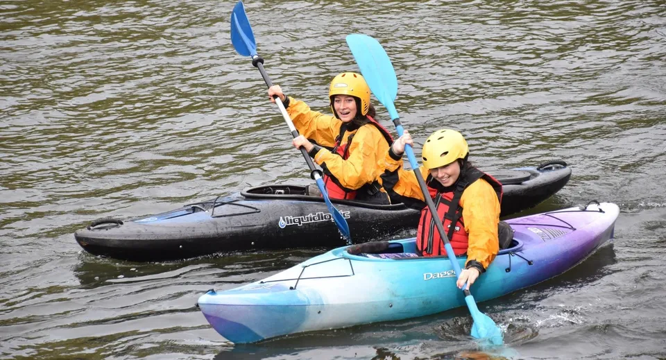 Un gruppo di studenti del Newstead College pagaia su un fiume in kayak colorati lungo una sponda alberata.