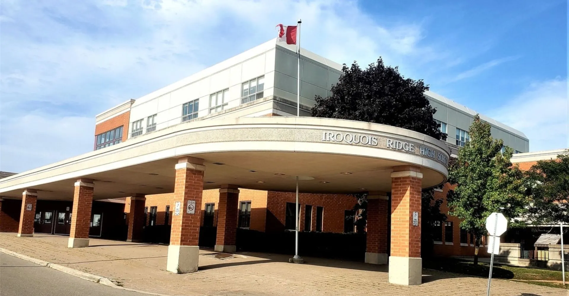 L'edificio principale moderno della Iroquois Ridge High School con la bandiera canadese sul tetto e alberi in primo piano.