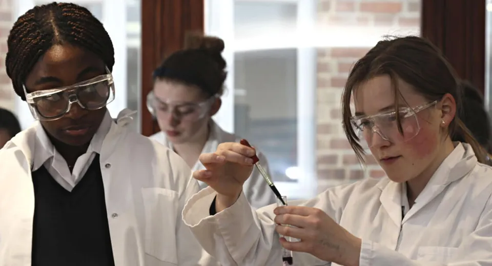 Tre studenti della Woldingham School lavorano concentrati in laboratorio con camici bianchi e occhiali protettivi.