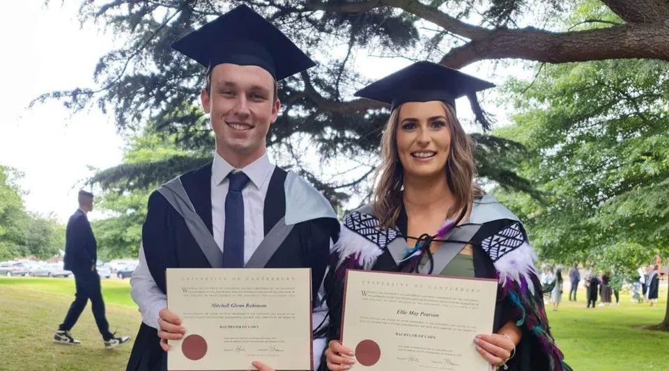 Due diplomati della Westland High School in toga tengono in mano i loro diplomi in un'area simile a un parco.