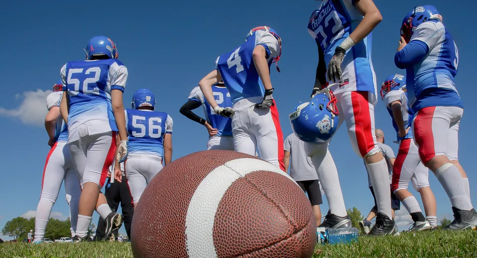 Un gruppo di giocatori di football della Collège Churchill High School si trova sul campo in erba con maglie blu e bianche e un pallone da football in primo piano.