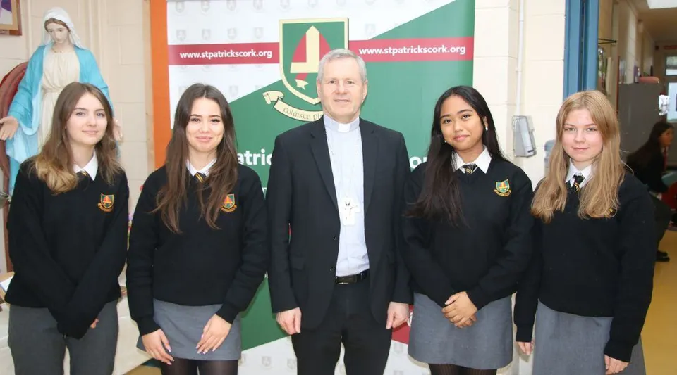 Un gruppo di studentesse e studenti del St. Patricks College in divisa scolastica si trova con un uomo anziano davanti a uno striscione verde e rosso.