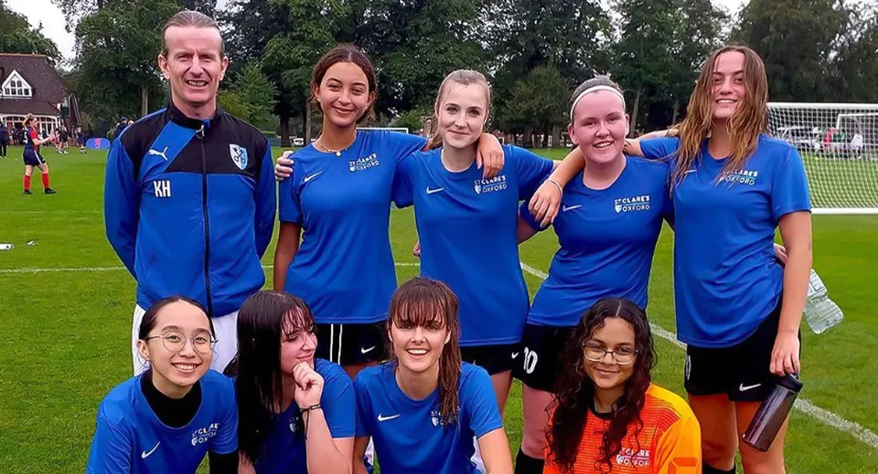 Un gruppo di studentesse della St. Clare’s School in tuta sportiva blu sta in piedi e seduto su un campo sportivo circondato da erba e alberi.