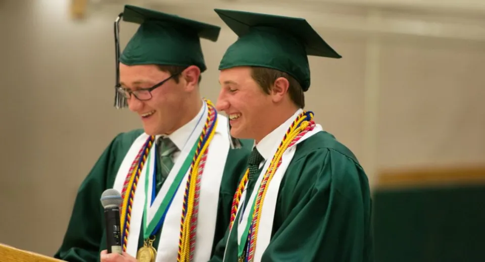 Due uomini in toga da laurea verde sono insieme e sorridono sul campus della Earl of March Secondary School.