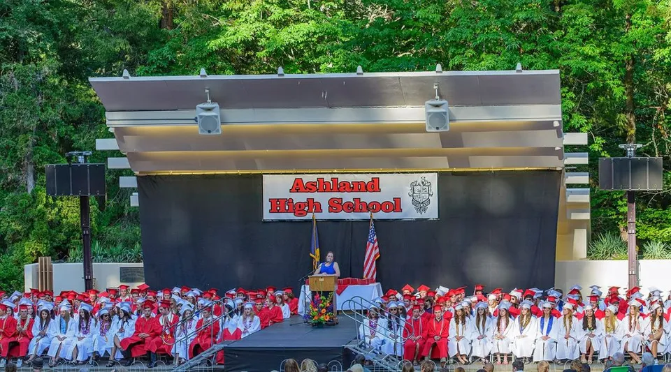 Studenti dell'Ashland School District indossano toghe rosse da diploma davanti a un palco con uno sfondo di bosco.