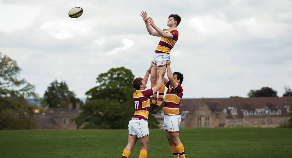 Due studenti della Downside School giocano a rugby su un campo sportivo con alberi sullo sfondo.
