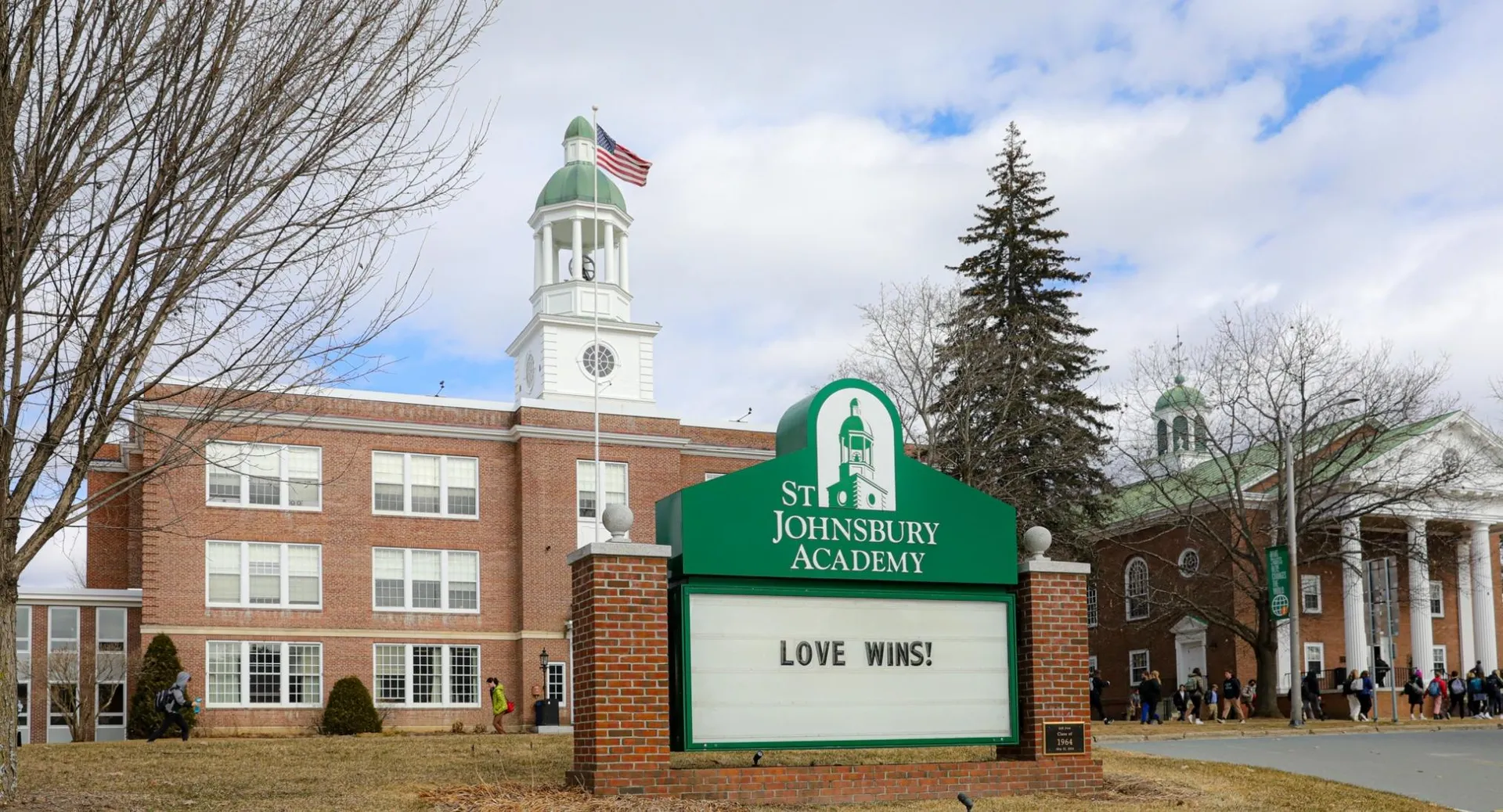 L'edificio in mattoni con torre dell'orologio e bandiera americana nel campus della St. Johnsbury Academy, circondato da alberi e prato.