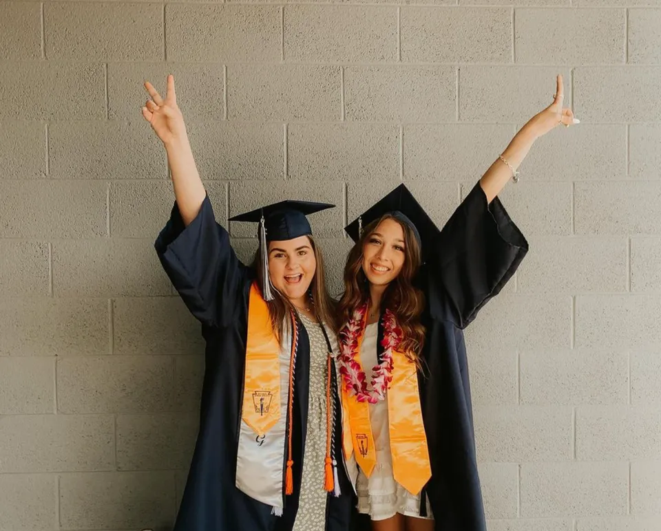 Due studentesse della Higley High School festeggiano il diploma con le braccia alzate davanti a una parete neutra.