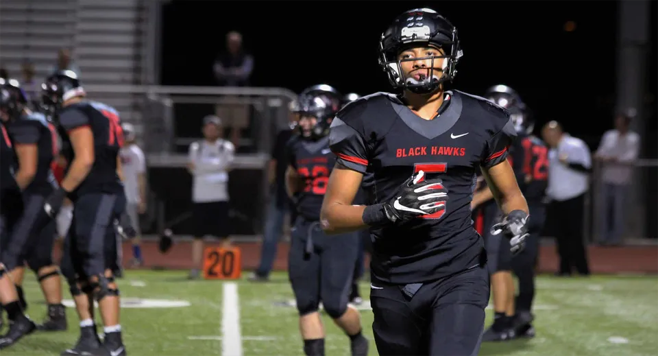 Un giocatore di football in uniforme nera e rossa è in piedi sul campo del Higley Unified School District, con altri giocatori e spettatori sullo sfondo.