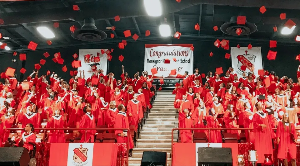 Studenti della Monsignor McCoy High School indossano toghe rosse durante la cerimonia di diploma in un ambiente interno.