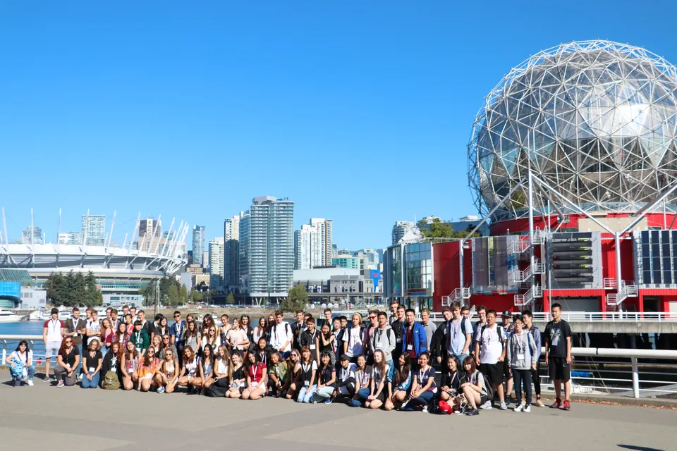 Un gruppo di studenti e studentesse si raccoglie davanti al moderno edificio a cupola della Burnsview Secondary School con il paesaggio urbano sullo sfondo.