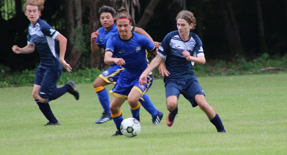 Studenti del Nambour State College giocano a calcio su un campo in erba con alberi sullo sfondo.