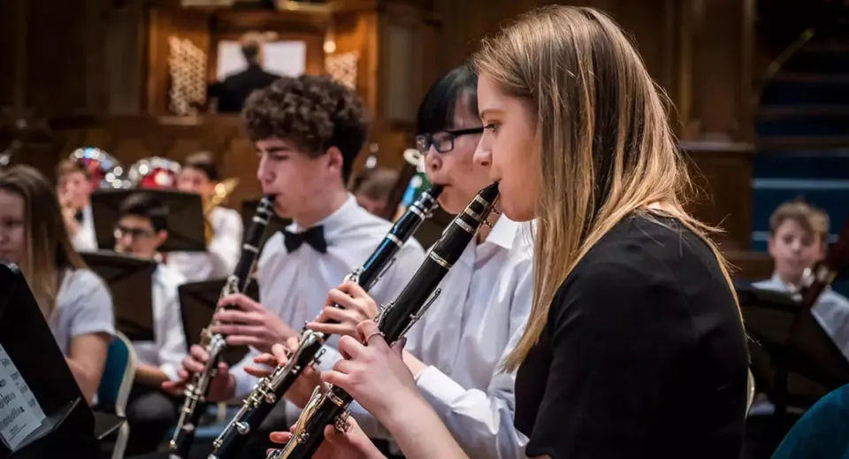 Un gruppo di giovani musicisti suona strumenti a fiato al Trent College davanti a uno sfondo di legno.