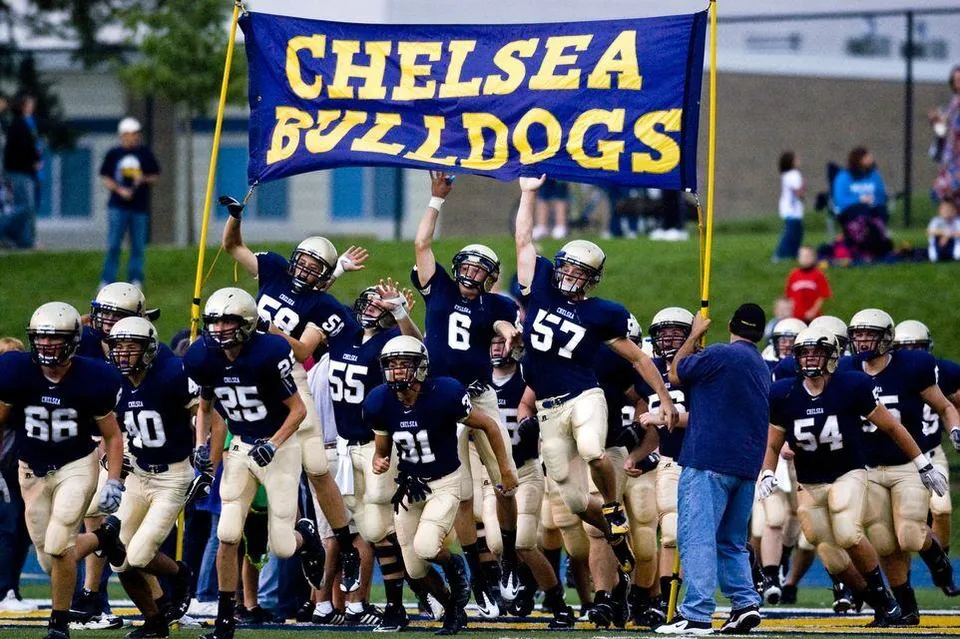 Studenti della Chelsea High School in uniforme da football sul campo di gioco con spettatori sullo sfondo.