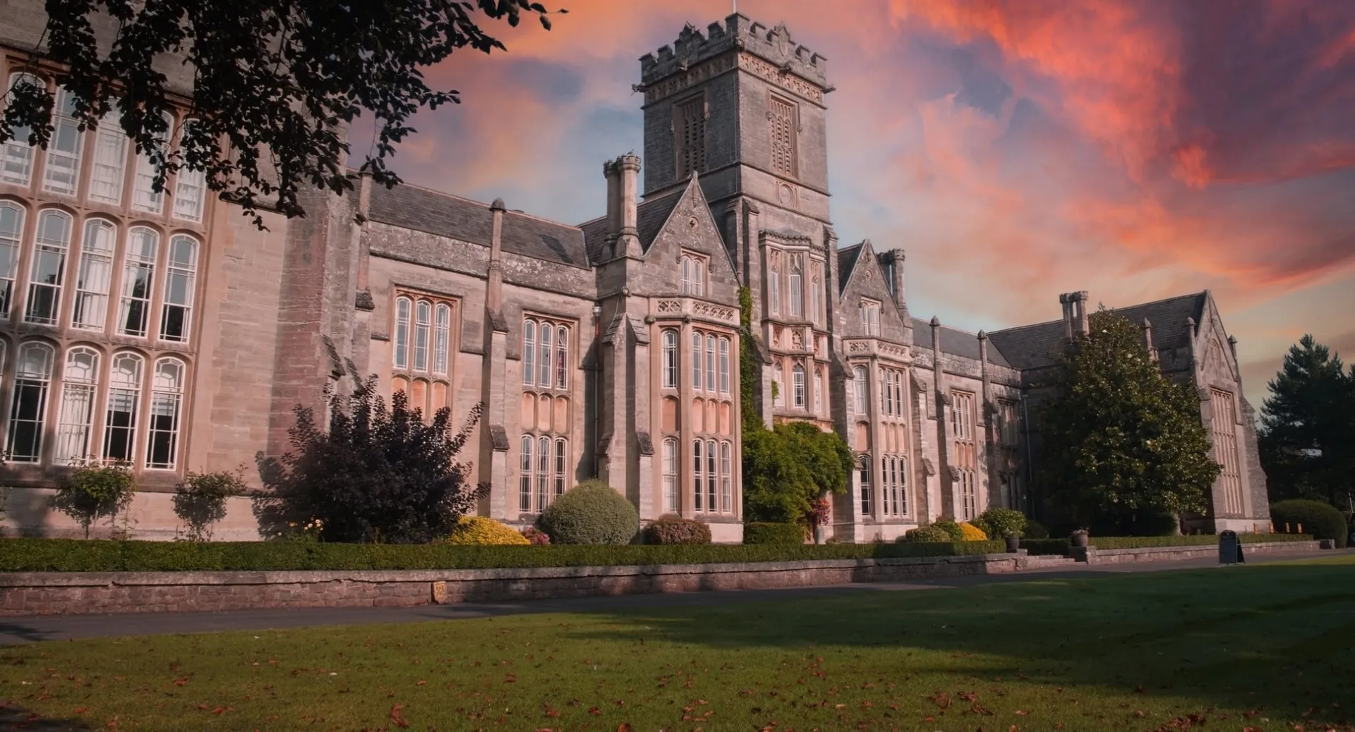 L'imponente edificio principale in pietra del Queen’s College è immerso nella natura verdeggiante e si staglia contro un cielo colorato al tramonto.