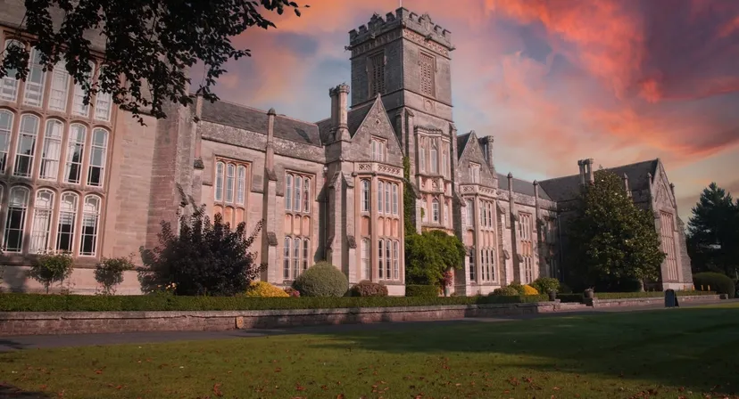 L'imponente edificio principale in pietra del Queen’s College è immerso nella natura verdeggiante e si staglia contro un cielo colorato al tramonto.