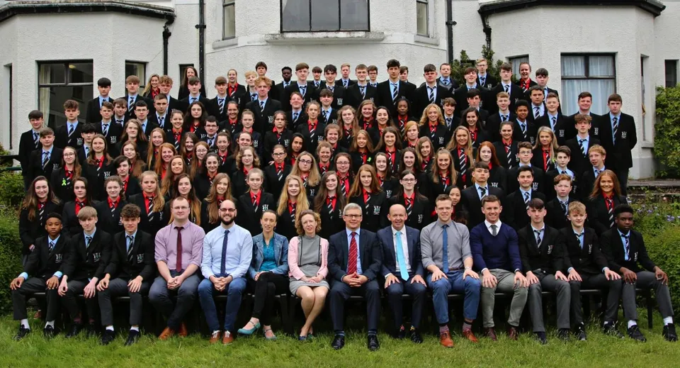 Un grande gruppo di studenti e studentesse in abito formale davanti a un edificio della Bandon Grammar School dall'architettura classica.