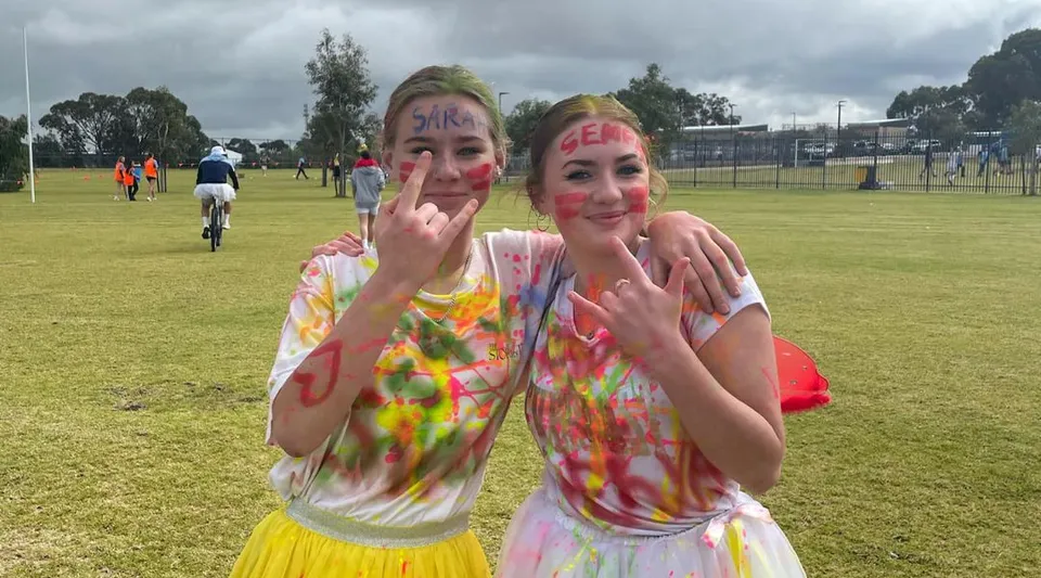 Due studentesse del Yanchep Secondary College indossano magliette colorate tie-dye e fanno smorfie all'aperto con gesti delle mani.