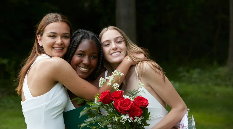 Tre studentesse della Oldfields School si abbracciano e tengono insieme un mazzo di rose rosse davanti a un giardino verde.