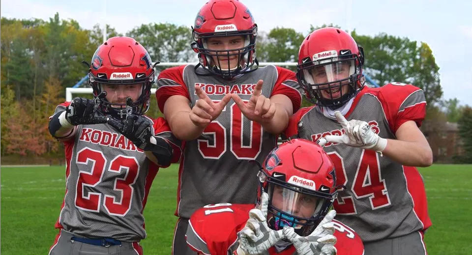 Quattro giocatori di football si trovano sul campo da gioco della Bear Creek Secondary School con alberi sullo sfondo.