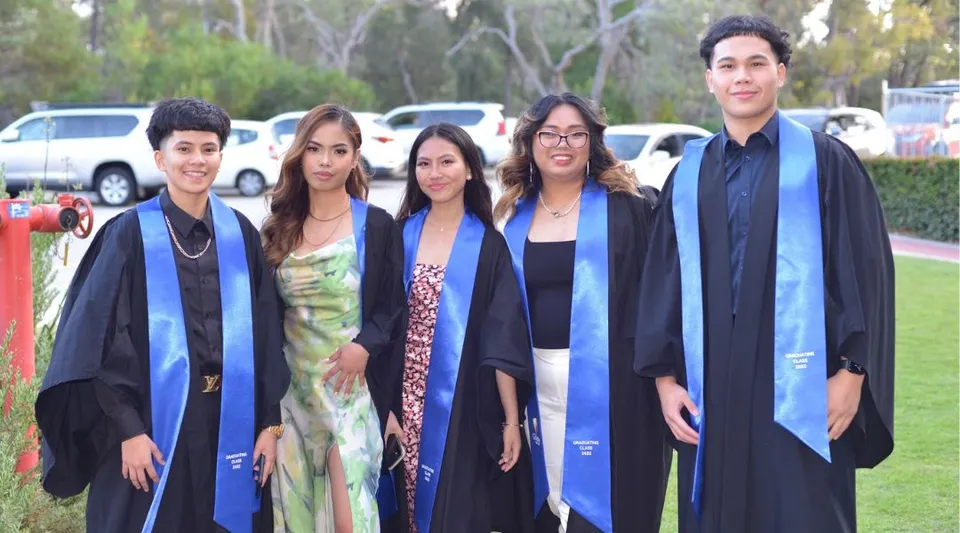 Un gruppo di cinque diplomati si trova all'aperto sul campus del Gilmore College indossando le toghe da diploma.