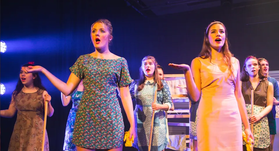 Un gruppo di studentesse della Burgess Hill Girls sta sul palco in abiti colorati in modo drammatico.