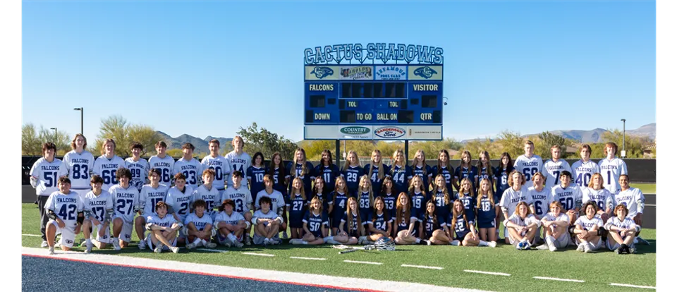 Giocatrici e giocatori di calcio della Cactus Shadows High School si trovano sul campo da gioco davanti a un tabellone segnapunti con le montagne sullo sfondo.