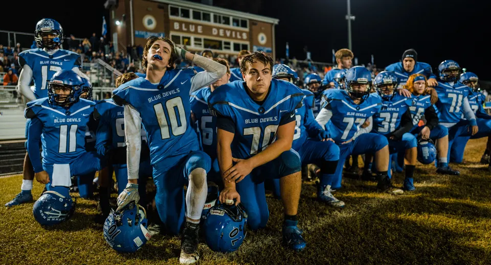 Un gruppo di giocatori di football in uniforme blu in piedi sul campo del Queensbury Union Free School District di notte.