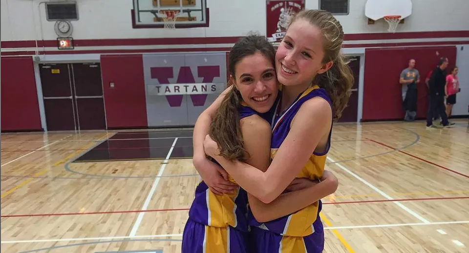Due studentesse della Wallaceburg District High School si abbracciano e sorridono sul campo da basket davanti a un cartello dei Tartans.