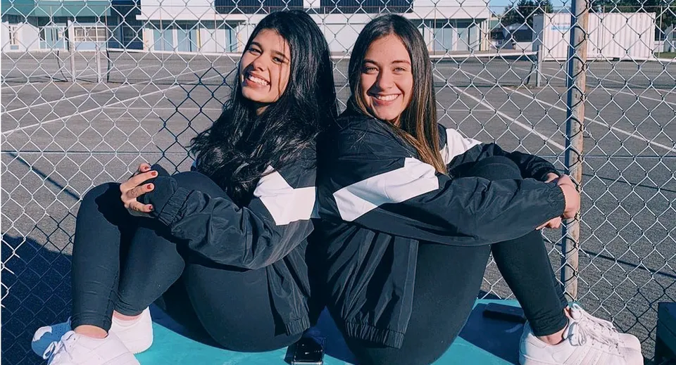 Due studentesse si abbracciano e sorridono davanti a una recinzione metallica nel campus della Sands Secondary School.