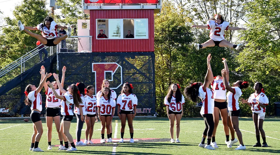 Studenti della The Pennington School eseguono acrobazie da cheerleader su un campo sportivo davanti a un edificio rosso e bianco.