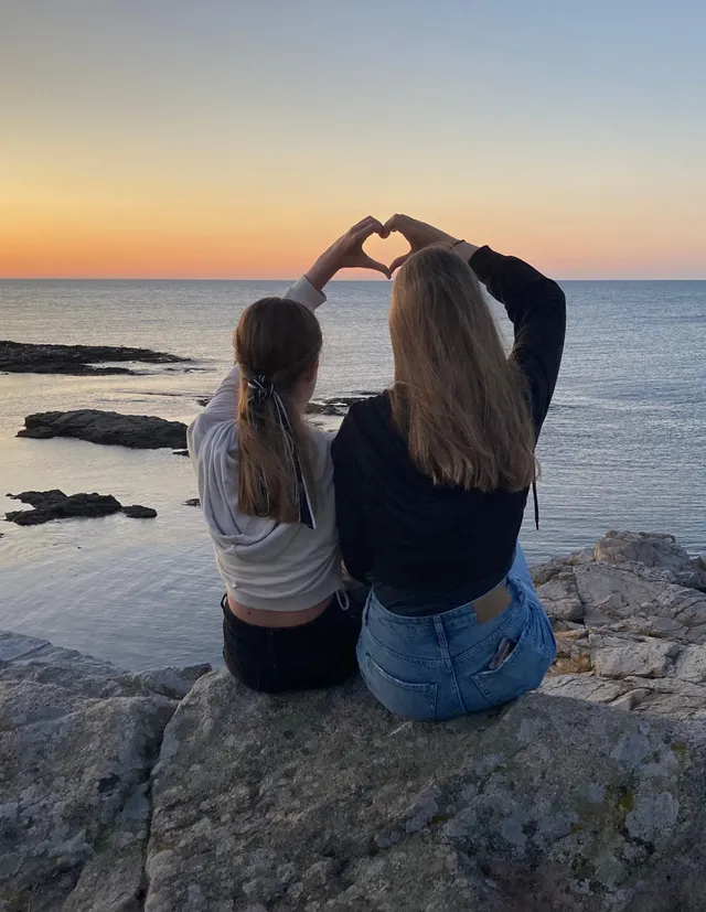 Dos chicas sentadas en las rocas formando un corazón con las manos frente al mar al atardecer