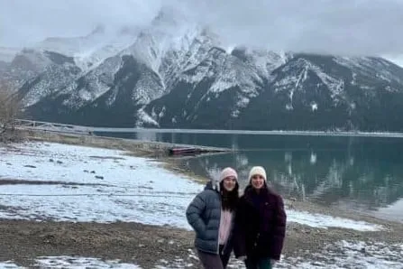 Dos chicas con abrigos de invierno frente a un lago y grandes montañas nevadas