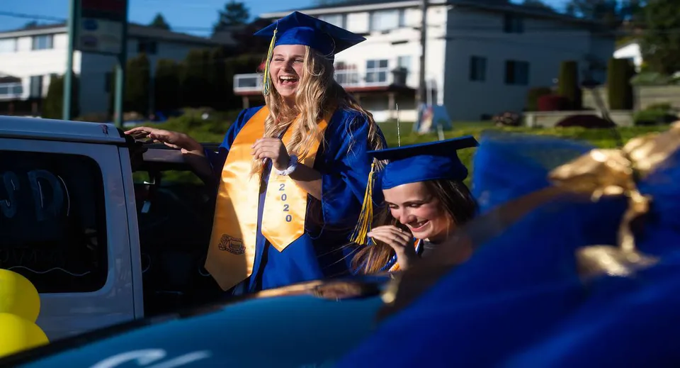 Una neolaureata sorridente in toga blu è accanto a un'auto nel campus del Torrance Unified School District, insieme ad altri neodiplomati.