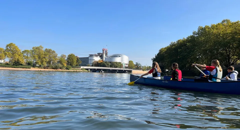 Un gruppo di studenti del Collège Episcopal Saint Étienne naviga su una barca blu su uno specchio d'acqua davanti all'edificio scolastico.