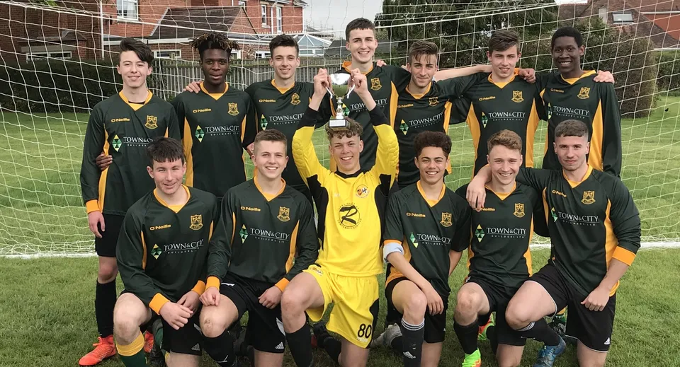 Un gruppo di studenti della St. Peter’s High School posa con un trofeo di calcio sul campo davanti a un edificio in mattoni.