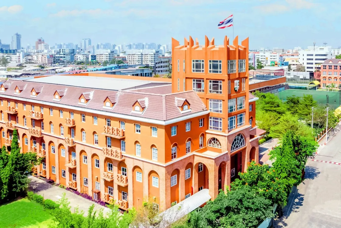 Luftaufnahme eines orangefarbenen Internatsgebäudes mit Turm und thailändischer Flagge, umgeben von Sportplätzen und Stadtsilhouette