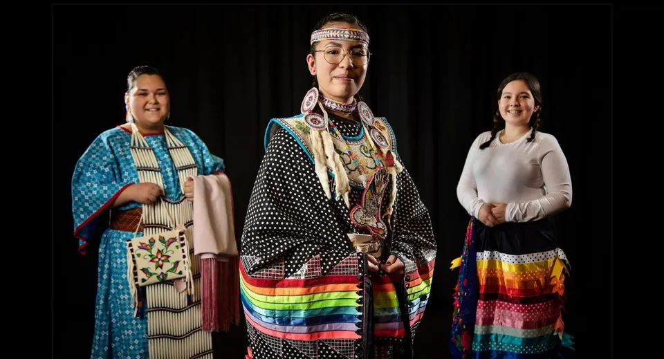 Tre persone in abiti tradizionali nativi americani davanti a uno sfondo scuro nel campus della Luther College High School.